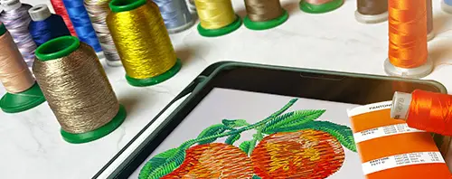 A digital tablet displaying an embroidered fruit design surrounded by colorful embroidery threads and a Pantone color guide, showcasing Dhaga-Raga’s creativity and attention to detail in custom embroidery design