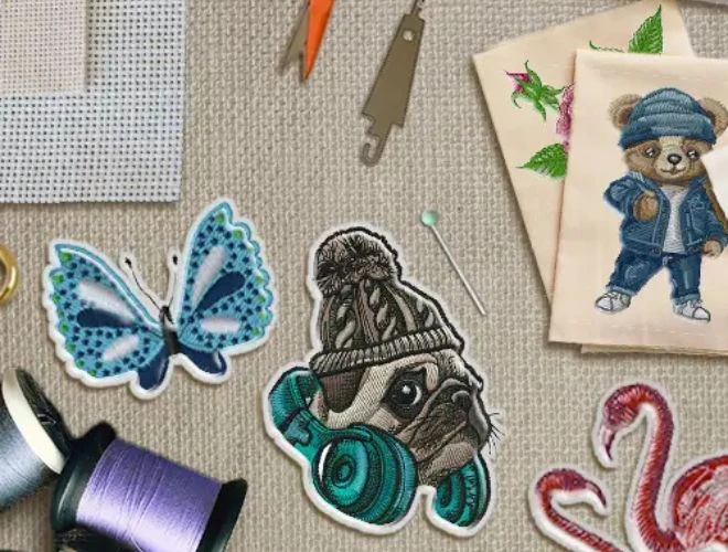 A flat lay of colorful embroidered patches including a butterfly, pug with headphones, flamingos, and a bear in denim, surrounded by embroidery tools and threads—highlighting Dhaga-Raga’s personalized embroidery services for any design or idea
