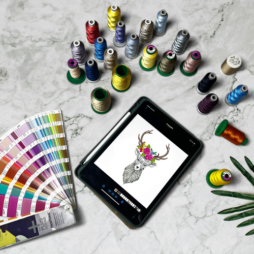 Embroidery design workspace featuring colorful thread spools, Pantone color guide, and a digital tablet displaying a floral antler deer artwork on a marble surface that is to be personalised or customised