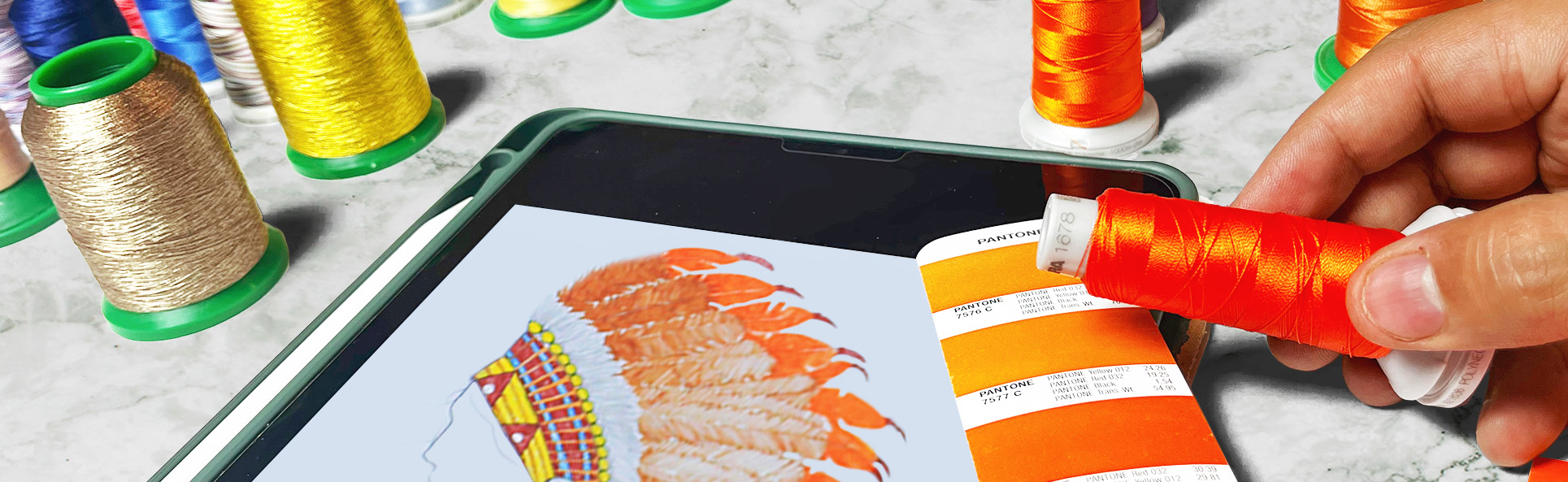 Hand selecting orange embroidery thread matching Pantone color swatches next to a tablet displaying a feather embroidery design