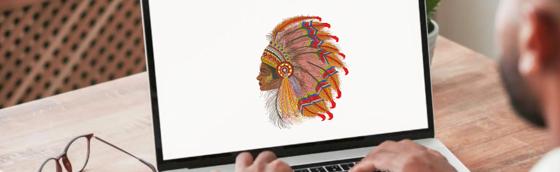 Person working on a laptop screen displaying a detailed embroidered design of a tribal headdress, showcasing digital embroidery artwork
